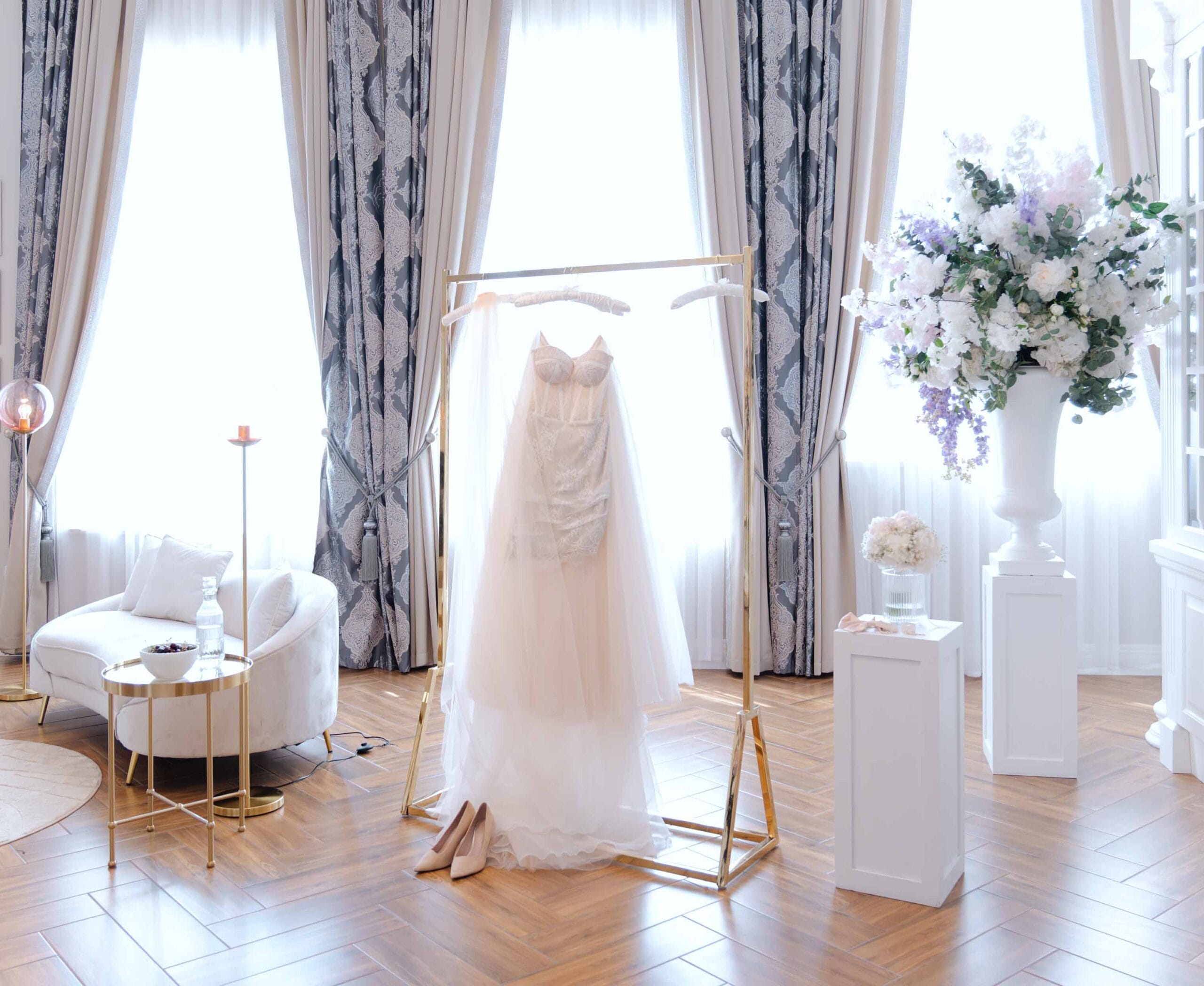 Elegancka suknia ślubna na wieszaku w jasnym, stylowym wnętrzu – przygotowania panny młodej, bukiet kwiatów, buty i romantyczna aranżacja sali, Weddatelier - Fotografia i Film Ślubny