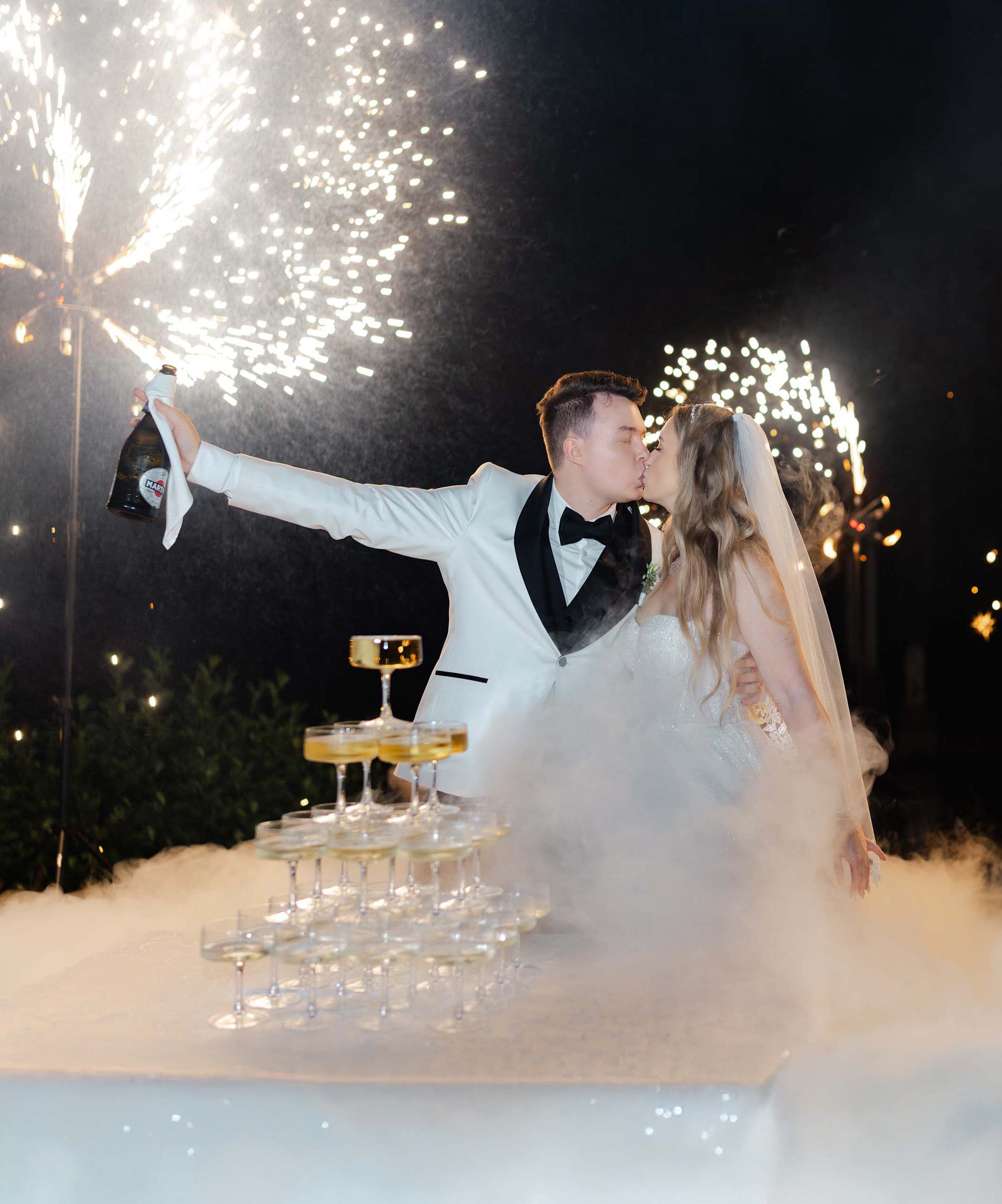 Nowożeńcy stoją przy eleganckiej wieży z szampana (Champagne Tower), otoczeni gęstym ciężkim dymem oraz pokazem efektownych fajerwerków. Romantyczna i luksusowa atrakcja ślubna uchwycona na profesjonalnej fotografii Weddatelier - Fotografia i Film Ślubny