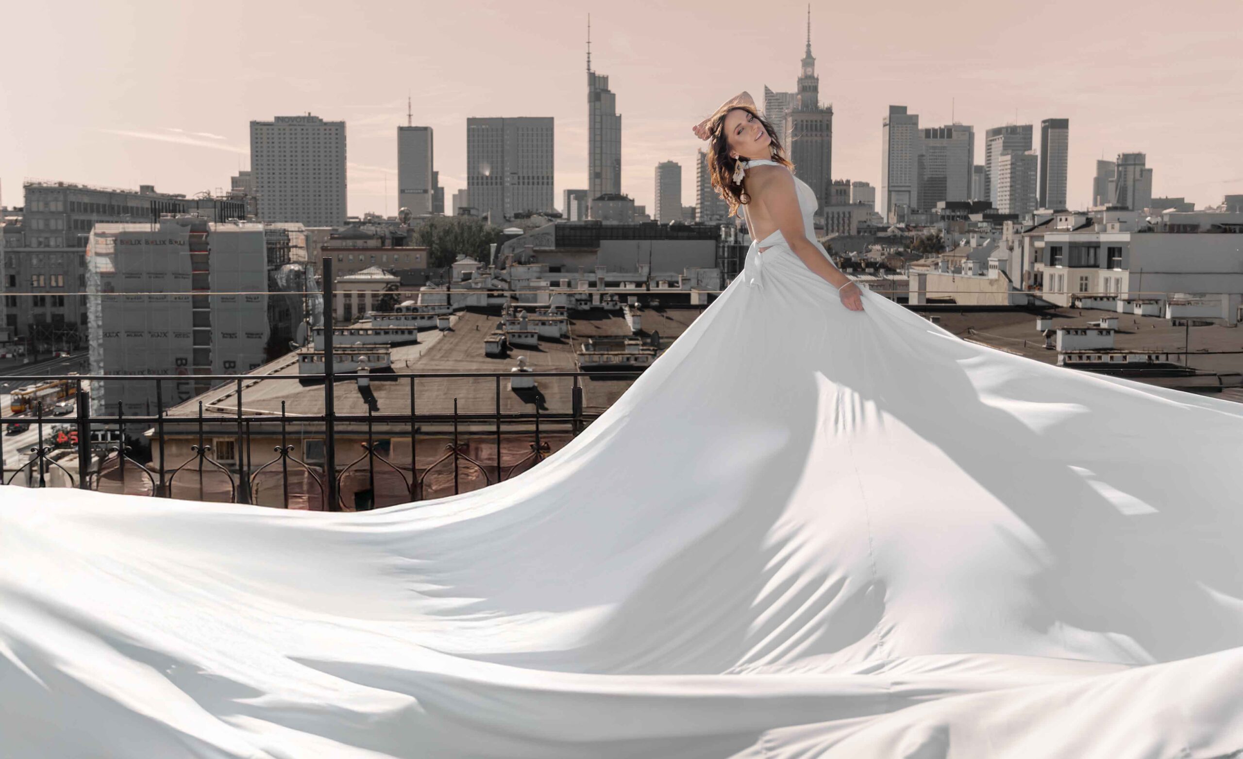 Sesja zdjęciowa w sukniach Flying Dress w Warszawie, modelka w długiej białej sukni, w tle panorama Warszawy Fotograf Ślubny Warszawa WeddAtelier.pl Fotografia i Film Ślubny