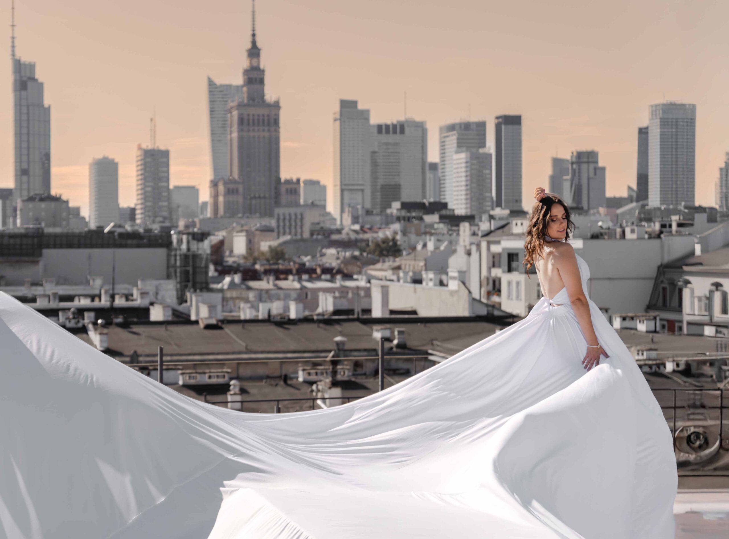 Sesja zdjęciowa w sukniach Flying Dress w Warszawie, modelka w długiej białej sukni, w tle panorama Warszawy Fotograf Ślubny Warszawa WeddAtelier.pl Fotografia i Film Ślubny