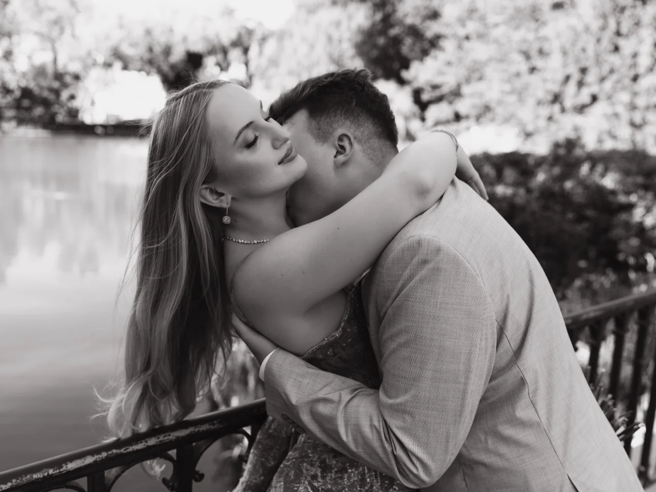 Fotografia ślubna Warszawa – WeddAtelier. Czarno-białe zdjęcie pary w intymnym uścisku przy kutej, żeliwnej balustradzie nad wodą. Mężczyzna w garniturze całuje kobietę w szyję. Kobieta w sukience wykończonej piórami odchyla się do tyłu z rozwianymi włosami.