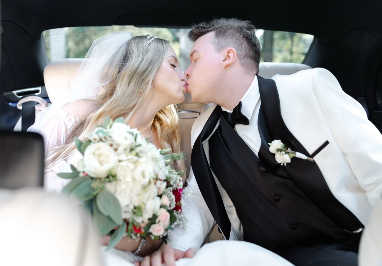 Fotografia ślubna Warszawa – WeddAtelier. Świeżo upieczeni małżonkowie całują się na tylnym siedzeniu luksusowego samochodu w drodze na wesele. Pan Młody ma na sobie elegancki, biały smoking z czarną muchą, a Panna Młoda ubrana jest w suknię z welonem, trzymając na kolanach jasny bukiet ślubny z zielonymi akcentami.