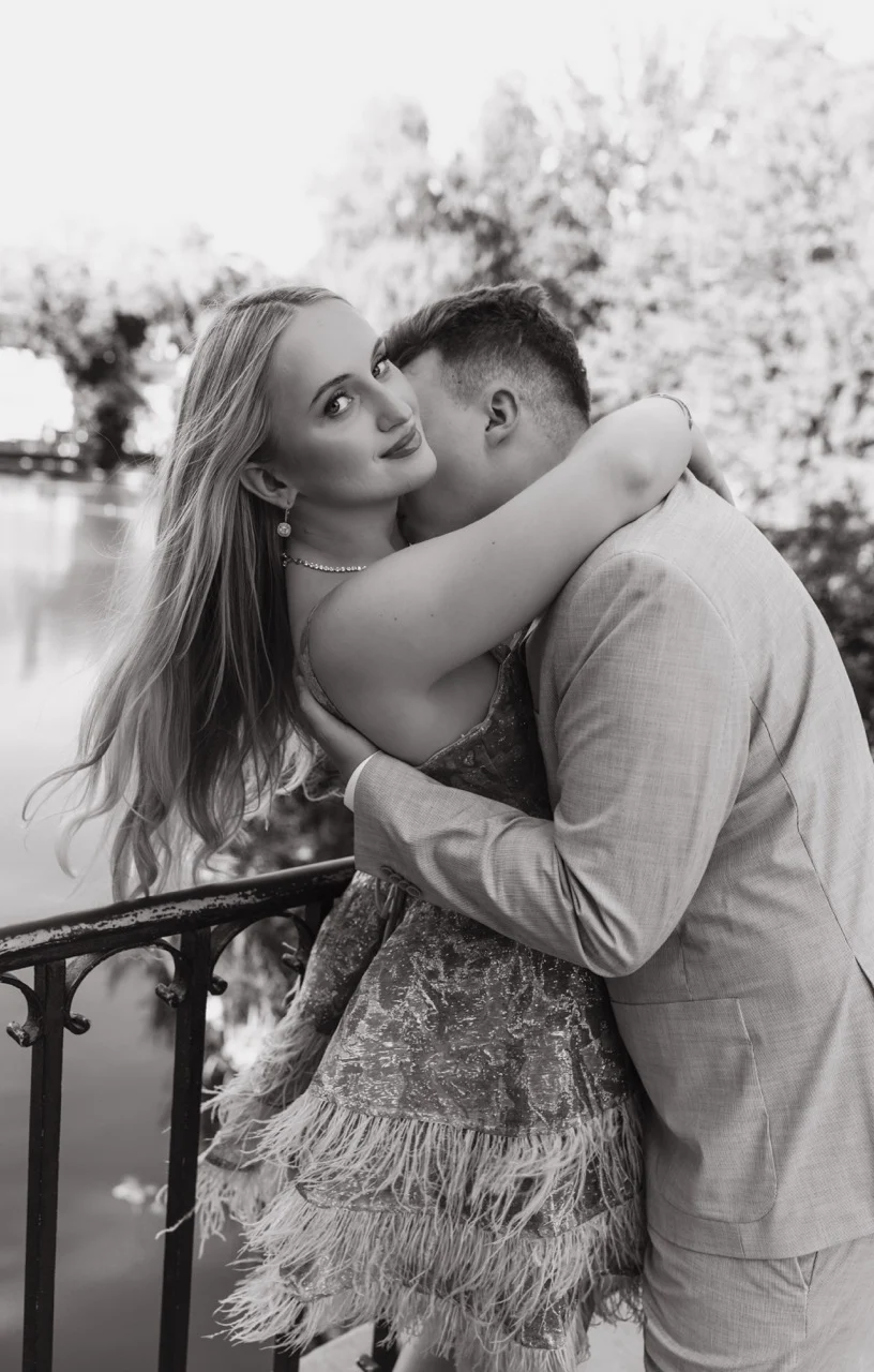 Fotografia ślubna Warszawa – WeddAtelier. Czarno-białe zdjęcie pary w intymnym uścisku przy kutej, żeliwnej balustradzie nad wodą. Mężczyzna w garniturze całuje kobietę w szyję. Kobieta w liliowej sukience wykończonej piórami odchyla się do tyłu z rozwianymi włosami, patrząc pewnym wzrokiem prosto w obiektyw aparatu.